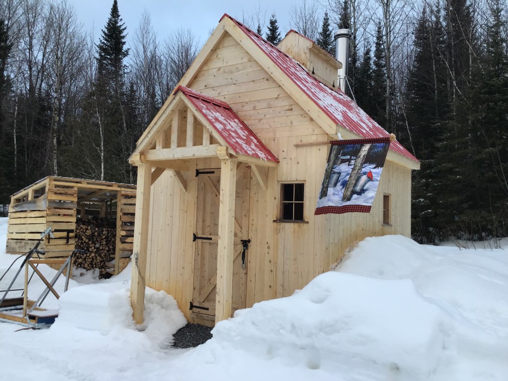 A small sugar shack with a red roof and woodshed beside it/Une petite Cabane à sucre aves un Toit rouge et un shed à bois à coté.
